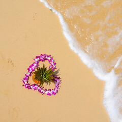 Heart-shaped flower lei around a golden pineapple at the beach with sand and waves