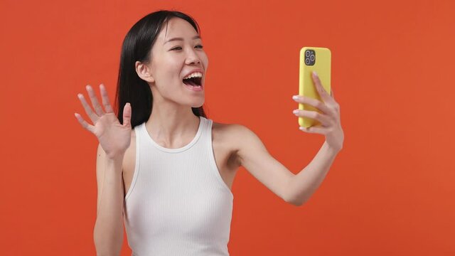 Young asian woman 20s years old wears white tank top shirt get video call using mobile cell phone doing selfie talk conducting pleasant conversation greet with hand on orange color background studio