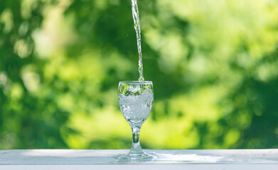 Water flows into a glass placed on a wooden bar,over sunlight and natural green background.blur...