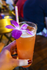 Vivid orange tropical cocktail with a purple orchid decoration with a ecological paper straw. Woman holding a cocktail glass by hand. Portrait mode photo. Tropical summer refreshment.