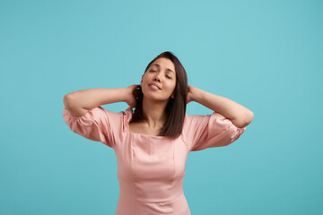 Fototapeta premium My hair like a silk. Relaxed carefree woman in pink dress keeps hands behind head, has positive expression, touched her hair, isolated over blue background. People expression concept.