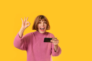 A girl stands in front of a yellow background and She plays with her cell phone She making "OK" action. She is very happy. Gamer girl is celebrating her victory making bet. She is smiling.