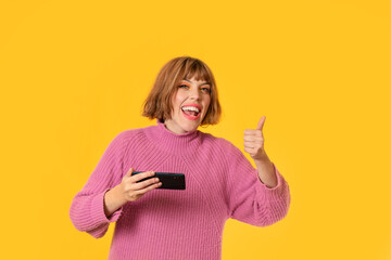 A girl stands in front of a yellow background and She plays with her cell phone She making "LIKE" action with her hand. She is very happy. Gamer girl is celebrating her victory making.