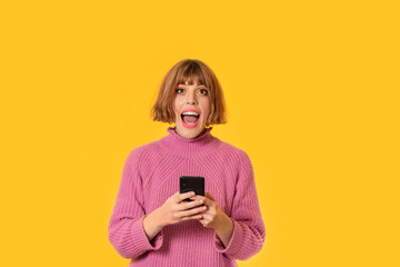 A girl stands in front of a yellow background and She plays with her cell phone. She looking at in front of. She is happy. Gamer girl is celebrating her victory making.