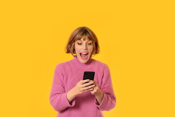 A girl stands in front of a yellow background and She plays with her cell phone. She looking at in hers telephone. She is very happy. Gamer girl is celebrating her victory making.