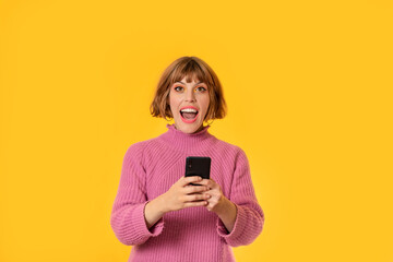 A girl stands in front of a yellow background and She plays with her cell phone. She looking at in front of. She is happy. Gamer girl is celebrating her victory making.
