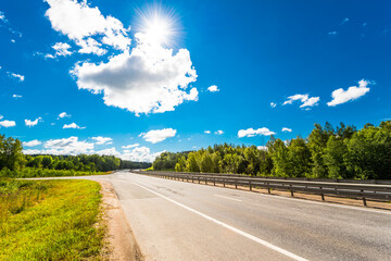 Rural road passing through fields and woods illuminated by the sun. View from the road