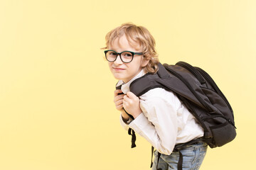 Sad schoolboy with glasses, with a large heavy backpack. Caucasian boy with blond hair on yellow...