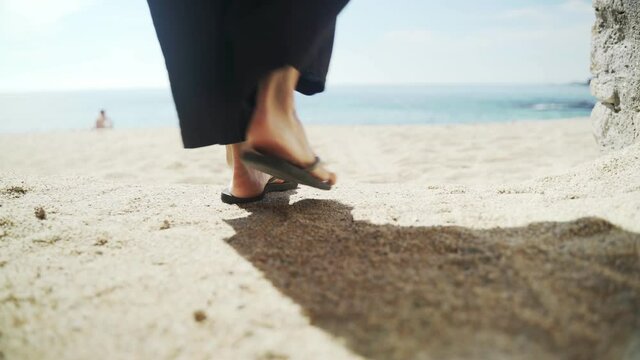 Young woman in wide black pants and a hat walks along the beach to the water. Takes off her black flip flops and goes barefoot. Sunny day at sea. Close-up of feet. Holidays on the beach. Summer chill