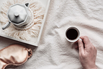 Espresso in hand, tray with white ceramic espresso coffee maker on trendy macrame pad. Sleep mask on crumpled bed sheets. Female hand with cup of espresso. Good morning Simple minimal flat lay.