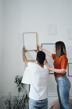 Excited african young husband and caucasian wife hang frame picture on wall happy settling together moving to new house, overjoyed biracial couple decorating own home relocating, ownership concept
