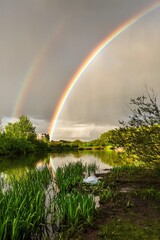 Vertical image of double colorful rainbow over grey sky and little pond with reflection in the...