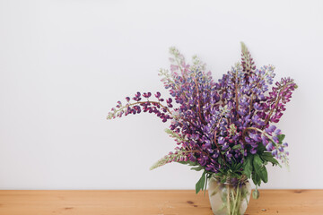 Beautiful lupine bouquet on wooden table in white room. Gathering countryside wildflowers, rural still life. Purple lupine flowers