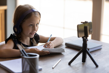 Happy little schoolgirl in headphones studying from home, using smartphone for watching online...