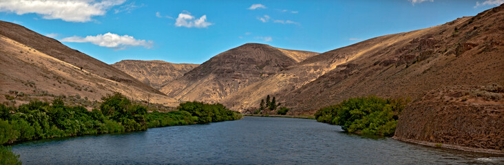 Yakima River Canyon in a panoramic view on a beautiful summer day with blue sky and a scattering of clouds and the river in the foreground. In central Washington.