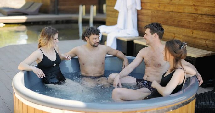 Company of friends bathing in outdoor jacuzzi or hot vat at the luxury spa. People relax have fun talking in wellness center