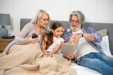 Obraz premium Small little girl granddaughter smiling when reading book with caring senior grandparents while lying on spacious bed enjoying leisure time with grandma and grandpa on weekend at home. Family concept