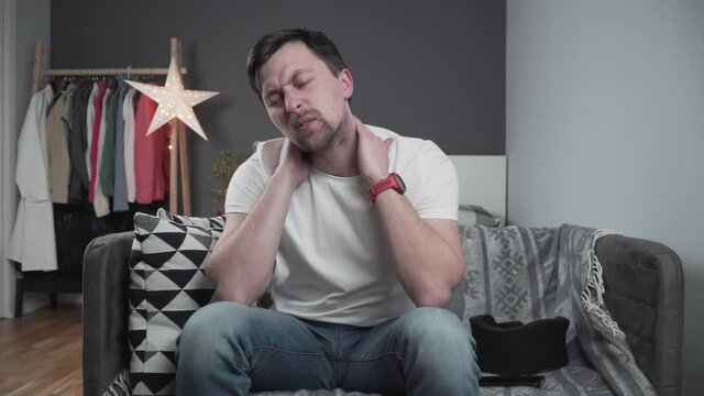 Guy wearing neck brace to support neck at home on couch. Caucasian man with neck injury uses collar support to relieve pain. Male in cervical brace relieves tension and pain in muscles of spinal