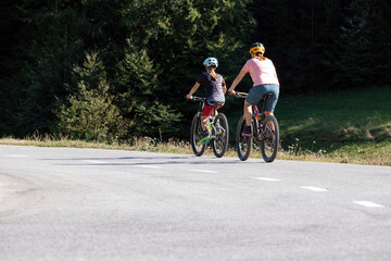 Naklejka premium Woman and girl child riding mountain bikes on a country road in nature on a bright sunny summer day.