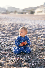 Small toddler in a blue overalls sits on his knees on a pebble beach and holds an apple. Close-up