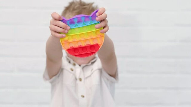 Portrait blonde boy with pop it sensory toy cat form. Boy presses colorful rainbow squishy soft silicone bubbles on white brick background. Stress and anxiety relief. Trendy fidgeting game.