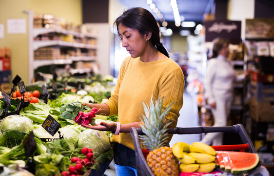 Portrait of pensive latin american woman buying fresh organic vegetables in supermarket, choosing red radish