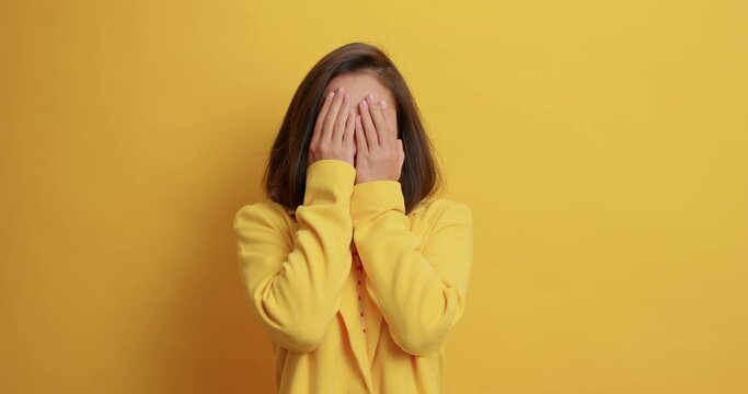 Scared frightened young Asian woman covers face with hands peeks through fingers witnesses terrible scene dressed formally isolated over yellow background. People emotions and reaction concept