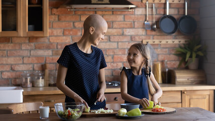 Happy ill mom and daughter kid talking and laughing while preparing salad and chopping fresh vegetables together. Cute girl helping mother with cancer to cook healthy dinner from organic food. - Powered by Adobe