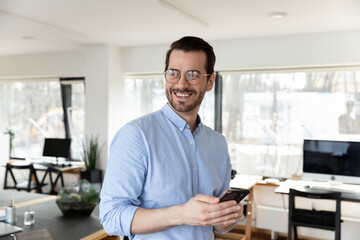 Happy dreamy businessman in glasses distracted from phone standing in office, looking to aside, smiling entrepreneur employee executive visualizing future, new opportunities, excited by good news