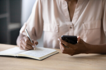 Close up young african ethnicity businesswoman holding cellphone digital gadget in hands, writing notes in paper planner, managing working assignments or web surfing important information online.