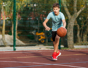 Obraz premium Cute boy in green t shirt plays basketball on a city playground. Active teen enjoying outdoor game with orange ball. Hobby, active lifestyle, sport for kids.