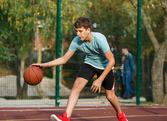 Fototapeta premium Cute boy in green t shirt plays basketball on a city playground. Active teen enjoying outdoor game with orange ball. Hobby, active lifestyle, sport for kids.