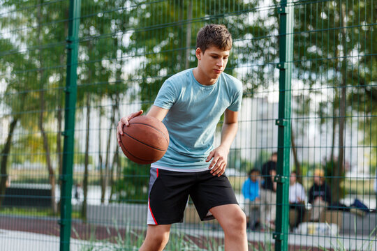 Cute boy in green t shirt plays basketball on a city playground. Active teen enjoying outdoor game with orange ball. Hobby, active lifestyle, sport for kids.