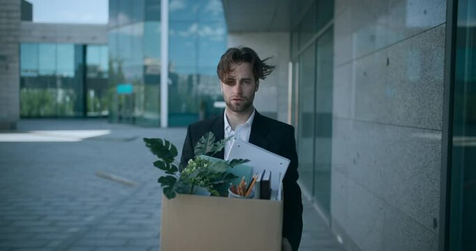 Outdoor portrait of depressed fired male employer walking out of office building with box of personal stuff