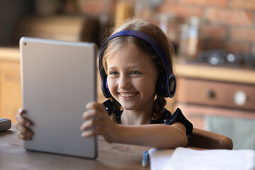 Happy little school girl in headphones using online app on tablet, talking via video call, watching movie or online lesson, sitting at table at hone, smiling at screen, Generation Z concept