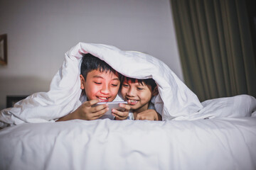 Happy asian brother playing online game on smart phone on the bed and  covering their body using blanket