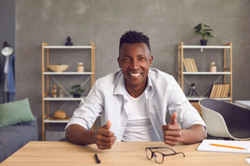 Happy friendly young African man sitting at table in home office. Web cam view of cheerful smiling black entrepreneur or college student. Webinar, videocall or videoconference laptop computer headshot
