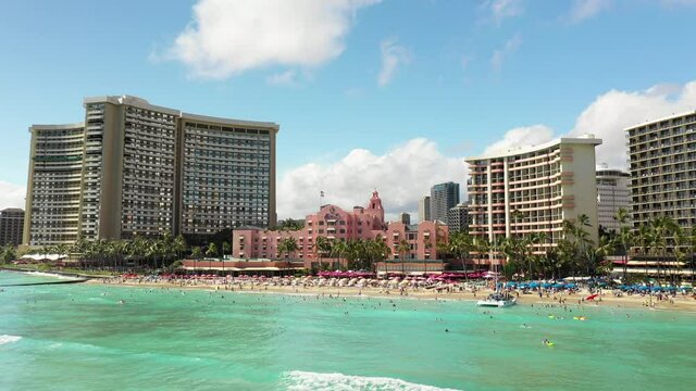Aerial Backward Shot Of Tall Buildings By Beach Against Cloudy Sky, Drone Flying Over People Surfing In Sea During Vacation - Waikiki, Hawaii