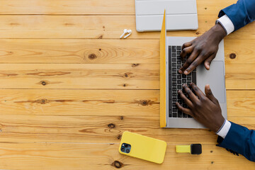 Top view of afro american men businessman or student hands working, typing with yellow laptop, smartphone and tablet at office wooden desk in modern office interior. Copy space.