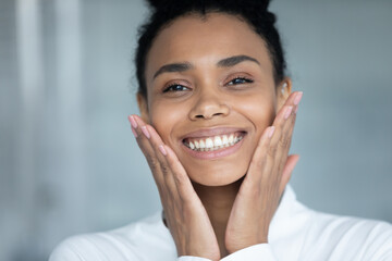 Head shot of happy joyful Black girl touching face in bathroom, admiring cosmetic effect of moisturizing cream, organic oil, cleaning lotion, looking at camera, smiling. Skincare, dental care concept