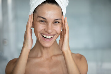 Head shot of happy woman with head wrapped in bath towel, touching face with toothy smile, applying...