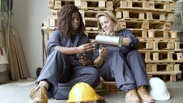 Happy female workers sitting at wooden platforms. Diverse women in uniform pouring coffee from thermos to mug, taking break from work and talking. Warehouse, coffee break concept.