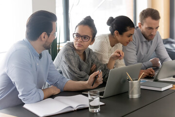 Diverse colleagues employees brainstorming, using laptops, working on project discussing strategy, sitting at table in boardroom, coworkers or student team sharing ideas, mentor training interns