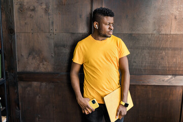 Serious focused afro american freelancer is standing near the wall, with yellow laptop, smart phone in hands, wearing casual trendy yellow and black outfit, looks far and misses someone