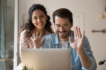 Happy candid family couple waving hand looking at laptop screen, starting online web camera video call conversation at home, talking speaking chatting remotely, distant communication concept.