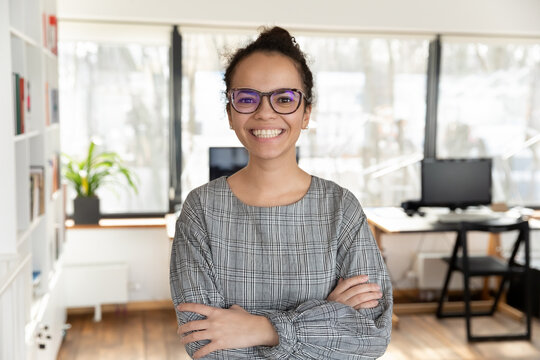 Head shot portrait of smiling African American businesswoman with arms crossed standing in modern office, happy confident successful executive employee intern woman in glasses looking at camera