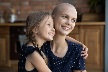 Positive daughter girl hugging happy ill young mom. Optimistic mother with cancer and kid enjoying leisure time together, embracing, looking away with home, thinking over good future. Oncology concept