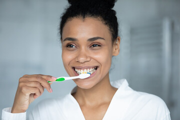 Happy teen African American girl in bathrobe cleaning and protecting white teeth with brush at...