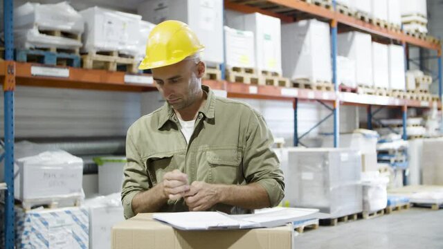 Focused male worker in helmet signing documents. Caucasian man checking order, examining box. Industrial labor, paperwork concept.