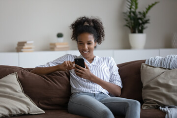 Addicted to modern technology millennial generation african mixed race woman relaxing on cozy sofa with smartphone in hands, involved in online shopping or distant communicating in social network.